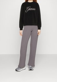 Svart sweatshirt med "Guess"-logotyp, ribbade muddar; grå ribbade byxor, utsvängd silhuett; grå sneakers med vita sulor.