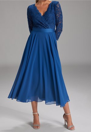 Blå kjole med blonder og lange ærmer samt en skålsyet halsudskæring, der har en tætsiddende overdel og en flydende nederdel. Sølvfarvede hæle med stropper er synlige.