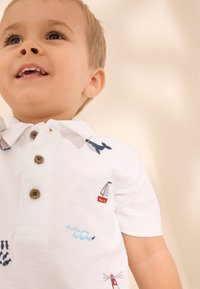 Weißes Polohemd mit gestickten Motiven eines Wals, Segelbooten, Wellen und eines Leuchtturms. Vier-Knopf-Verschluss am Kragen.