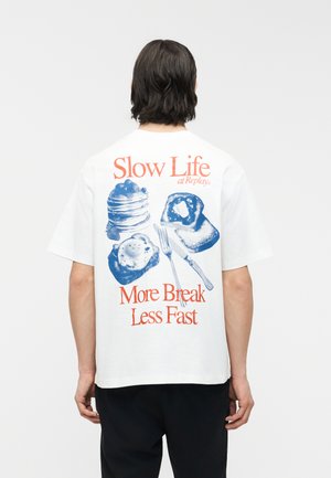 Person som har på seg hvit T-skjorte med blått grafisk motiv av frokostting og oransje tekst som sier "Slow Life More Break Less Fast" på baksiden.