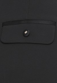 Tejido negro con una textura suave que presenta un bolsillo con solapa y un botón redondo negro con un sutil acento metálico.