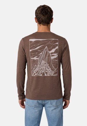 Hombre con camisa marrón de manga larga con un diseño en línea blanca de montaña y nube en la espalda, combinado con vaqueros azul claro.