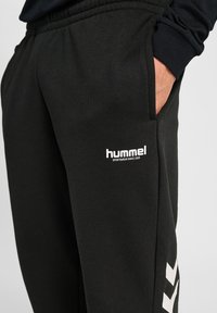 Zwarte sportbroeken gemaakt van zacht materiaal, met een elastische tailleband, zijzakken en een wit "hummel" logo aan de voorkant.