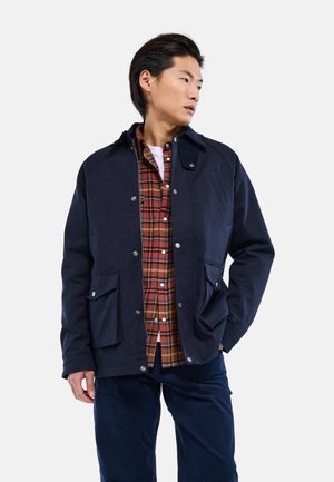 Homme portant une veste bleu marine par-dessus une chemise à carreaux rouge et orange boutonnée, avec un pantalon foncé, regardant vers sa gauche sur un fond uni.