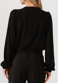 Zwart langslijf top gemaakt van gestructureerde gaasstof met een diamantpatroon, voorzien van puffed mouwen en een ontspannen pasvorm aan de achterkant.