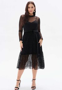 Czarna sukienka z dopasowanym gorsetem i przezroczystymi długimi rękawami. Warstwowa spódnica ma wzór w panterkę i ozdobny detal w postaci związania w talii.