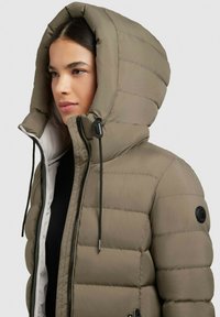 Padded parka jacket in olive green with a large, hooded collar. Features a black logo patch and adjustable drawstrings at the hood and zipper.