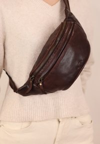 Marsupio in pelle marrone con due scomparti con cerniera, una texture liscia e un design curvo, indossato a tracolla sopra un maglione chiaro.