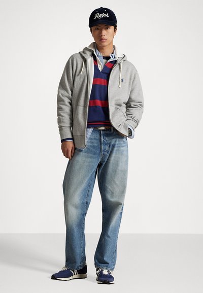 Grå hettejakke med glidelås over en navy- og rødstripet polo, blå jeans og navy sneakers. Svart caps med "Ralph"-logo. Uformell passform.