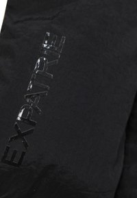 Sort stof med en struktureret overflade, der har det præget ord "EXPATRIÉ" i en blank finish. Designet er minimalistisk og moderne.