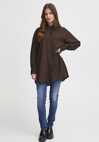 Loose-fitting brown shirt with a collar and button closure, paired with blue skinny jeans and black ankle boots, on a plain background.