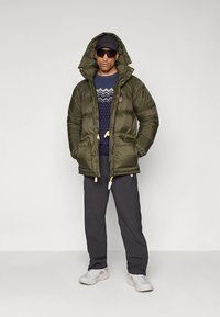 Groene pufferjas met capuchon, gele trekkoorden en dubbele zakken aan de voorkant, gedragen over een marineblauwe trui met patroon en donkere broek met grijze sneakers.