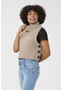 Beige, bordázott kötött ujjatlan pulóver magas gallérral és három sötét gombbal az oldalon, világoskék farmernadrággal párosítva.