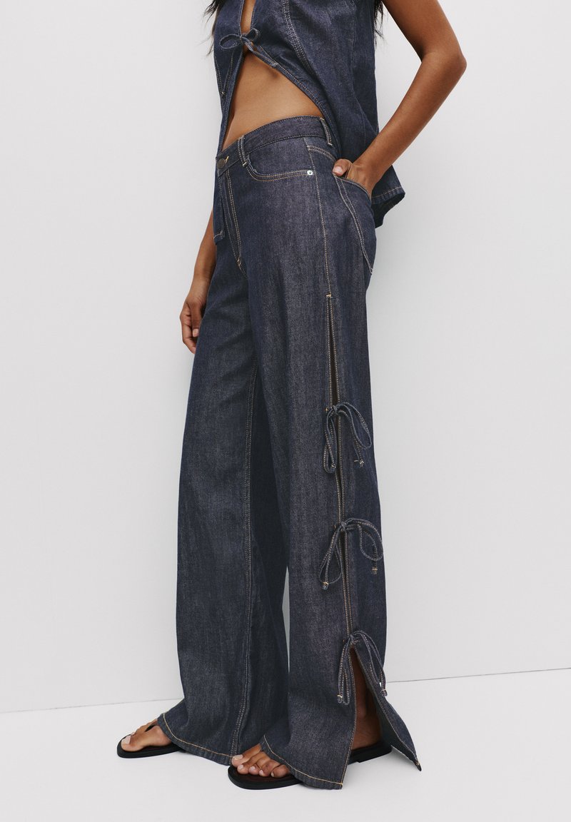 Mörkblå denim byxor med vida ben, sideluckor och knytband, med kontrasterande sömmar och hög midja.