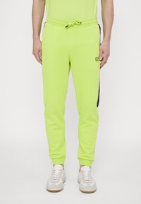 Neon zelené tepláky s elastickým pasem a šňůrkou. Obsahují boční černý pruh a detail loga. Měkká a hladká textilie.