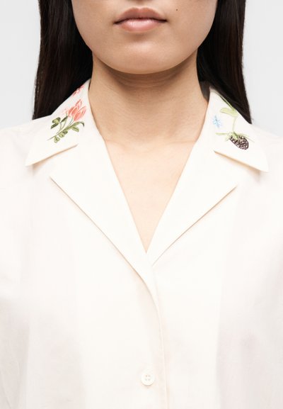 Close-up de uma pessoa vestindo uma camisa creme com padrões bordados de flores e bagas em cada ponta da gola.