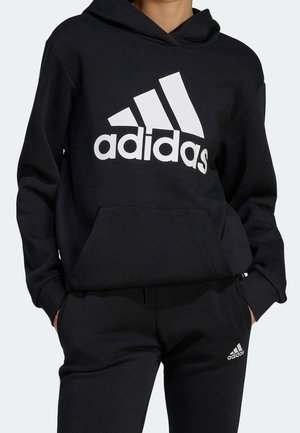 Person trägt schwarzen Adidas-Hoodie mit großem Logo auf der Brust und schwarze Adidas-Sweathose mit Logo am linken Oberschenkel. Hände in den Taschen.
