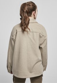 Béžová oversized košile s měkkou texturou, s špičatým límcem, spadlými rameny a dlouhými rukávy. Zadní strana má jednoduchý design bez zdobení.