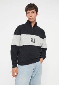 Svetrový ovládací blok v námorníckej modrej a sivej farbe s vysokým golierom, disponujúci predným zipsom a vyšitým logom "GAP". Mäkkučká látka, voľný strih.