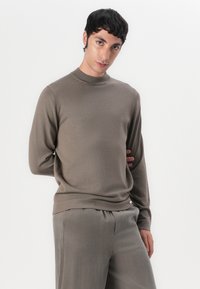 Maglione a maglia marrone chiaro con collo alto e maniche lunghe, abbinato a pantaloni leggeri dello stesso colore, con una texture liscia.