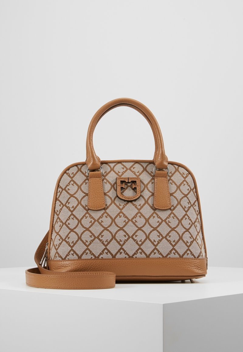 Sac à main à motifs marron et beige avec deux poignées, bandoulière amovible et un emblème de logo central sur un piédestal blanc.