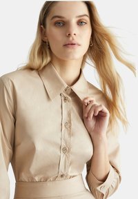 Camicia di raso beige con colletto appuntito, maniche lunghe e bottoni in pietra decorativa lungo la parte anteriore. Tessuto liscio e design aderente.