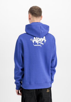Man facing away wearing a blue hoodie with white "Alpha Industries" logo on the back and black pants with a yellow patch on the pocket.