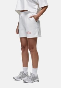 Shorts bianchi di cotone con tasche laterali e stampa grafica rosa, abbinati a sneakers grigie e calzini bianchi alla caviglia.