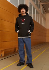 Svart huvtröja med en röd och vit Chicago Bulls-logga, tillsammans med blåjeans och svarta sneakers. Har randiga ärmslut och fåll.