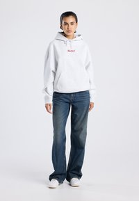 Mujer de pie con una sudadera gris clara con el texto "New York", jeans azul y zapatillas blancas, frente a un fondo claro y liso.