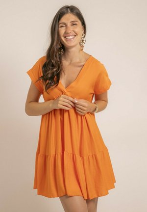 Femme souriante aux longs cheveux bruns, portant de grandes boucles d'oreilles en or et une robe orange à manches courtes avec un col en V, se tenant devant un fond uni.