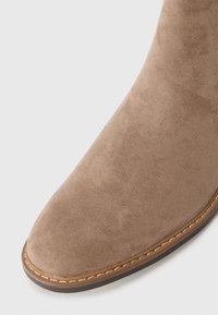 Bota de tobillo de ante marrón con punta redondeada, que presenta un ribete cosido y una textura suave. La vista en primer plano destaca el material y la forma.