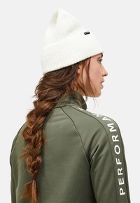 Groene prestatiejack met een wit zijlogo; gecombineerd met een geribde witte muts. Lange, gevlochten bruine haren zichtbaar vanaf de achterkant.