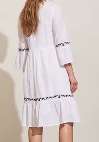 White cotton dress with black floral embroidery on sleeves and hem. Features a flared skirt and three-quarter bell sleeves. Textured fabric.