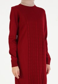 Vestido de punto rojo con un patrón de trenzas en el centro. Mangas largas y puños acanalados. La tela tiene una textura suave y una silueta ajustada.