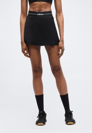 Αθλητική γυναίκα που φοράει μαύρη φούστα-παντελόνι Adidas, μαύρο αθλητικό σουτιέν, μαύρες κάλτσες και μαύρα παπούτσια, όρθια μπροστά σε απλό φόντο.