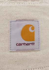 Štítek Carhartt na texturované béžové tkanině. Štítek obsahuje charakteristické oranžové logo vedle slova "carhartt" napsaného tmavě vínovým písmem.