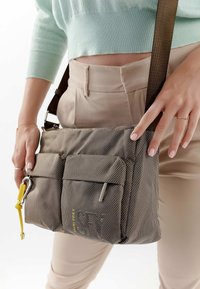 Donna con pantaloni beige e top corto verde menta che tiene in mano una borsa a tracolla grigia con texture, con tasche frontali e un portachiavi giallo.