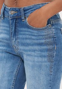 Blå denimjeans med en tettsittende design. Har en klassisk femlommers stil og er pyntet med dekorative nagler på den ene siden.