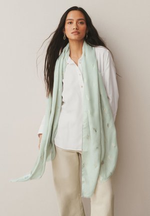 Mladá žena s dlhými tmavými vlasmi, nosiaca zlaté náušnice, biele tričko, béžové nohavice a svetlozelený šál s jemnými vzormi.