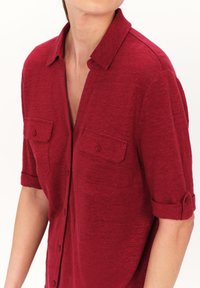 Camisa roja de manga corta con cuello, que presenta dos bolsillos en el pecho y mangas enrollables, hecha de un tejido texturizado. Cierre de botones en el frente.