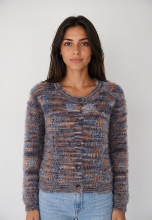 Donna con capelli lunghi e scuri che indossa un cardigan morbido multicolore con bottoni e jeans azzurri chiari, in piedi davanti a uno sfondo chiaro e semplice.