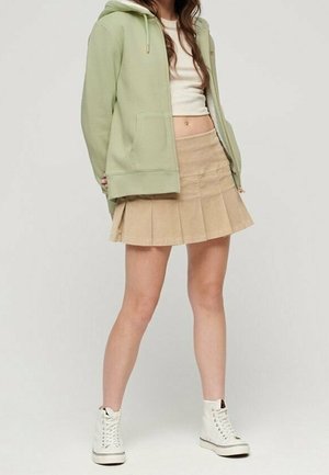 Personne portant un sweat à capuche zippé vert clair, un crop top blanc, une jupe plissée beige et des baskets montantes blanches, debout devant un fond uni.