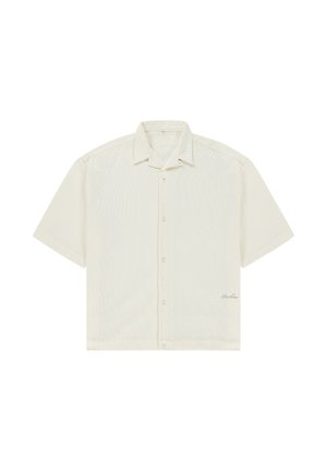 Camicia crema a maniche corte con bottoni, colletto e tessuto leggermente strutturato, con piccole scritte ricamate vicino all'orlo inferiore.