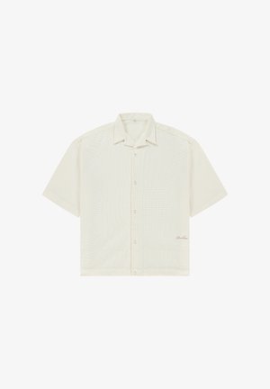 Kortærmet cremefarvet skjorte med knapper, krave og et subtilt tekstureret stof, med små broderede bogstaver nær nederste kant.