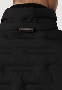 Chaqueta negra acolchada con un cuello alto, presenta un parche con un logotipo y una textura suave. El material parece ligero y aislante.