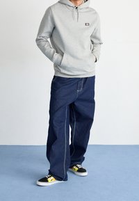 Persona che indossa una felpa grigio chiaro, jeans scuri larghi con cuciture bianche e scarpe nere-gialle, in piedi su un tappeto azzurro chiaro.