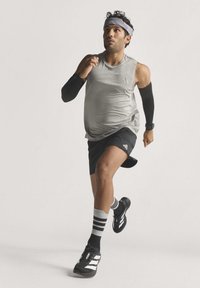 Mann, der ein graues ärmelloses Shirt, schwarze Shorts, ein Stirnband und schwarze Armsleeves trägt, läuft vor einem einfarbigen Hintergrund nach vorne.