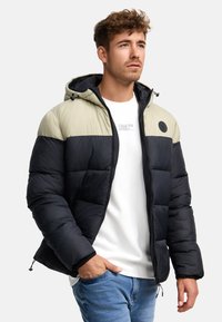 Mlad muškarac nosi beige i crnu puffer jaknu sa raznobojnim blokovima preko bijelog dukserice i plavih traperica, s jednom rukom u džepu.