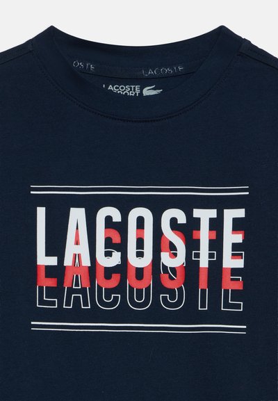 Тъмносиня тениска с кръгло деколте с думата "LACOSTE", отпечатана с големи бели, червени и очертани букви на гърдите.